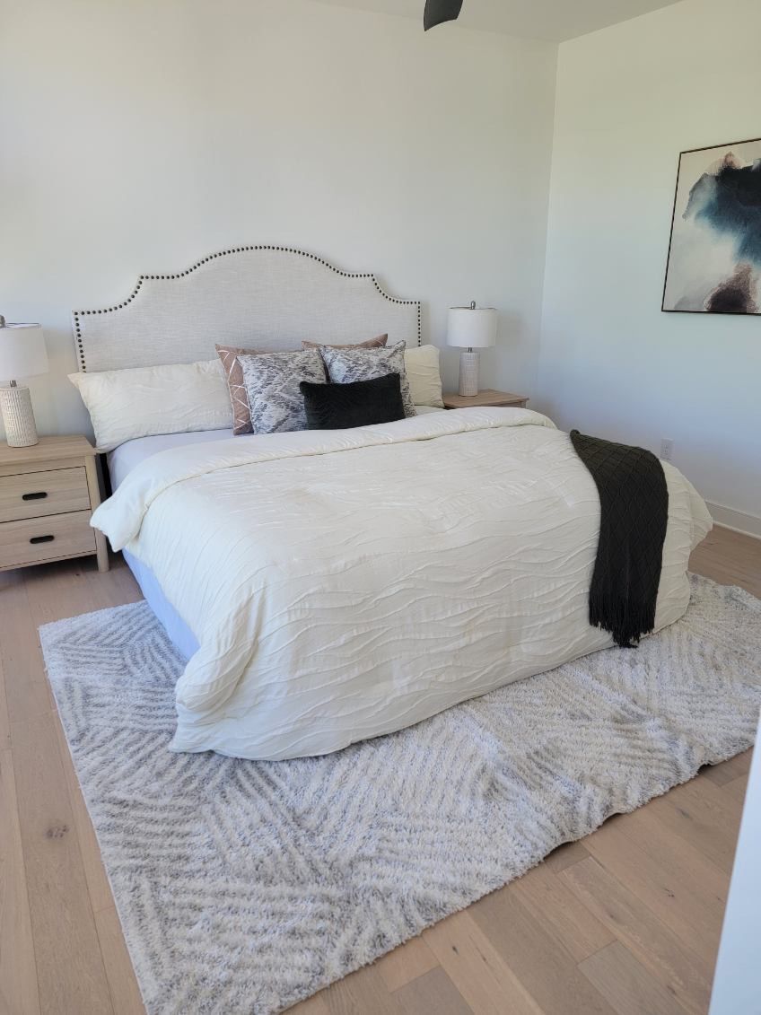 Bedroom with a large bed, white bedding, and a patterned rug on light wood floors.