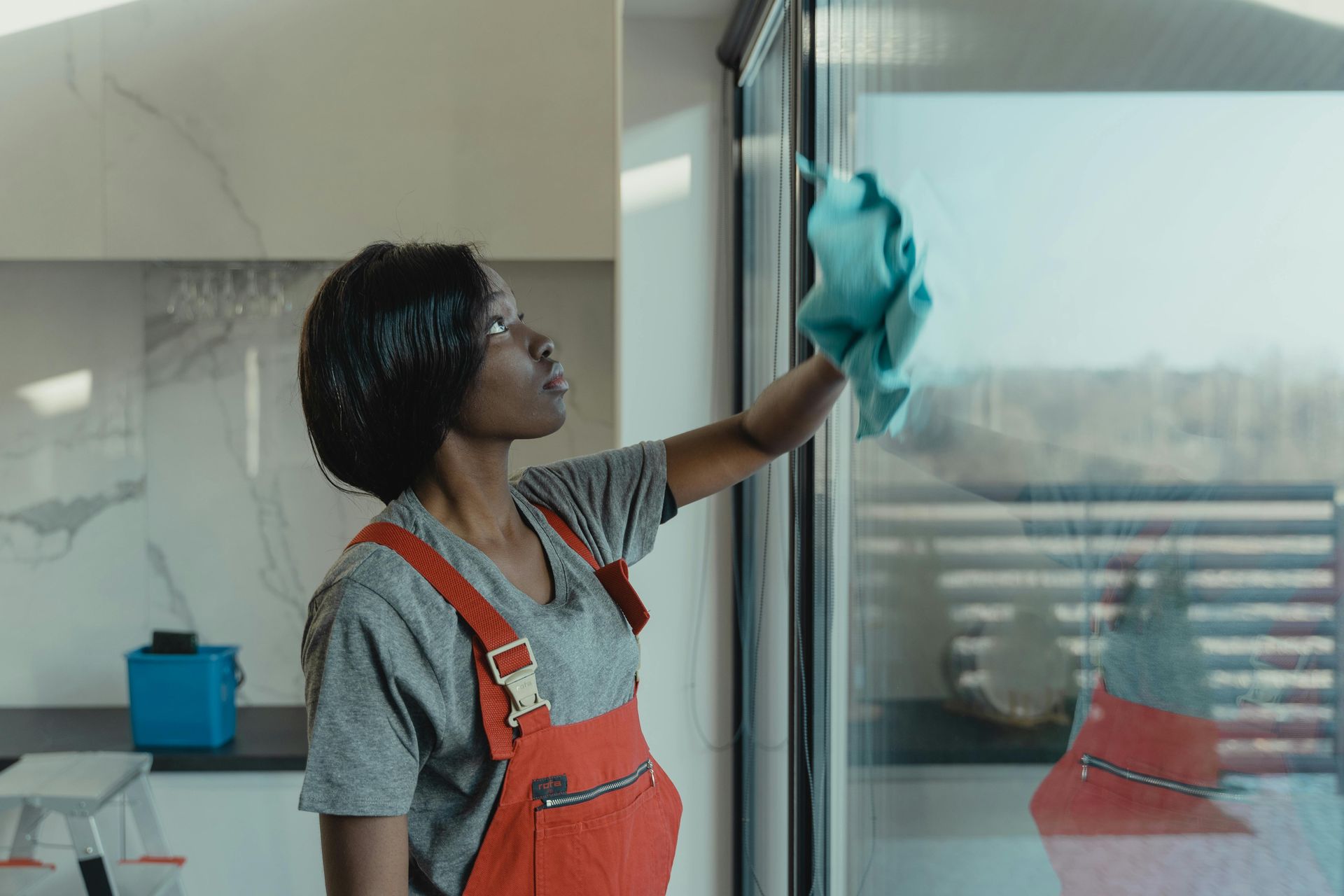 Woman in red overalls cleans a glass window with a blue cloth in a modern kitchen.