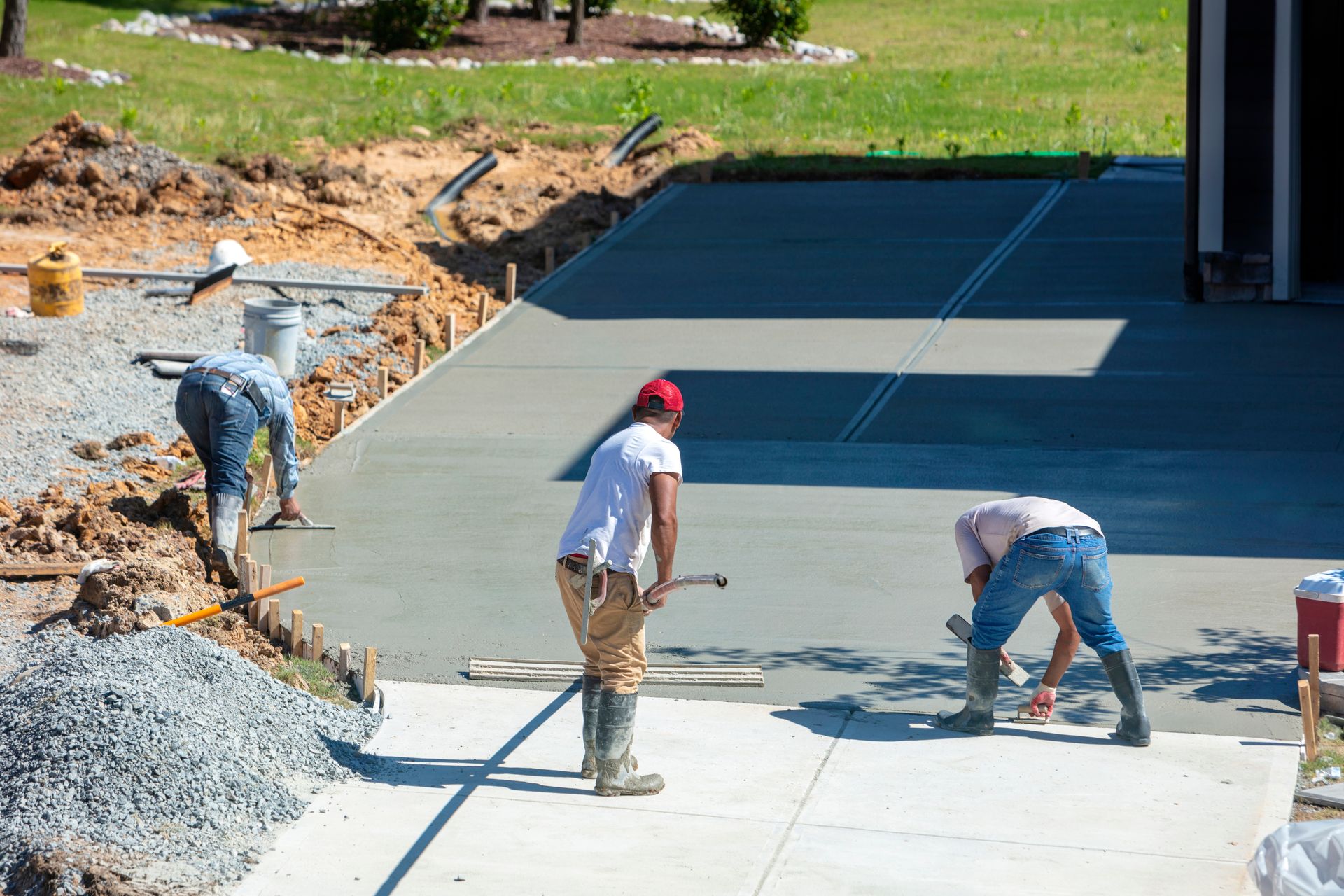 Three workers smoothing fresh concrete driveway. Gravel pile, building in background, sunny day.