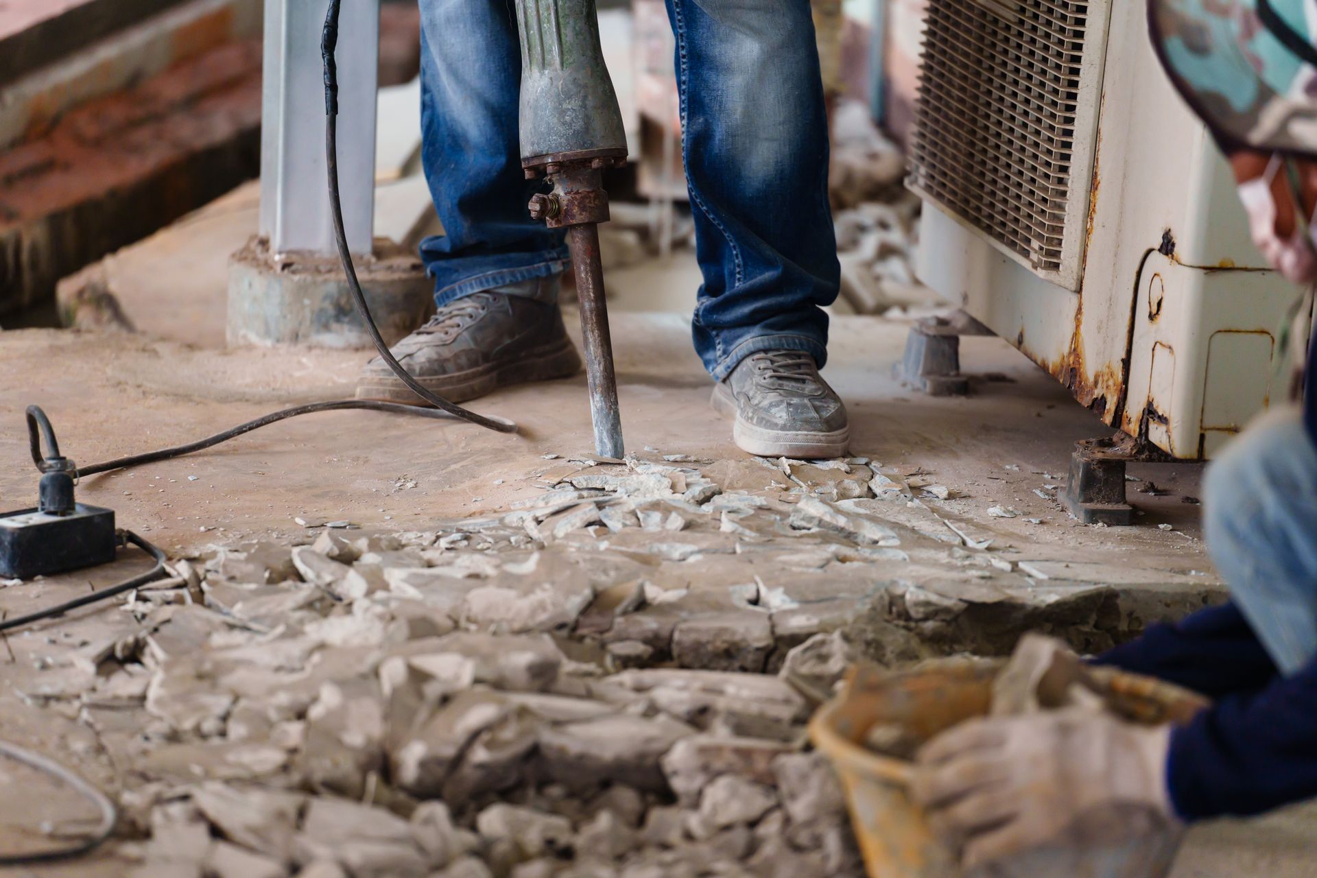 Construction worker using a jackhammer to break up concrete, another worker nearby with a bucket.