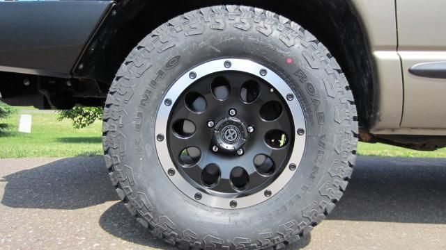 Close-up of a truck’s off-road tire and black rim on pavement, with the vehicle’s side visible.