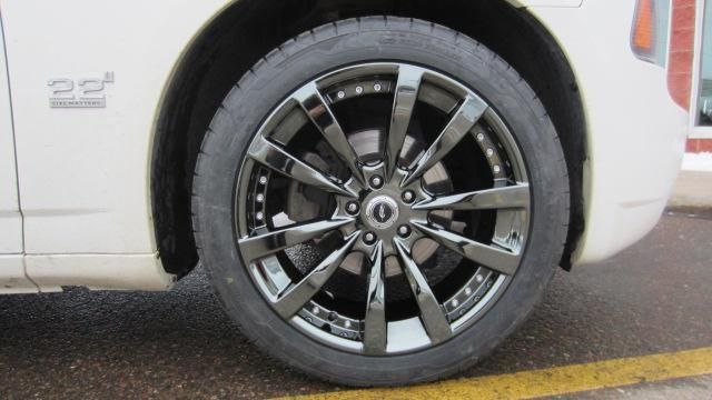 Close-up of a silver car’s rear wheel and tire parked on asphalt by a yellow line.