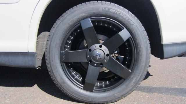 Black five-spoke car wheel and tire on a white vehicle parked on pavement