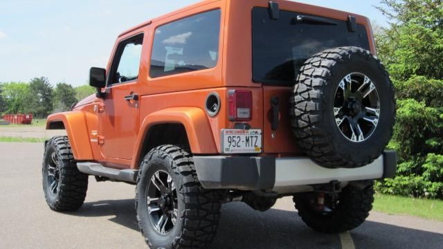 Orange Jeep Wrangler with large off-road tires parked on a road, rear view
