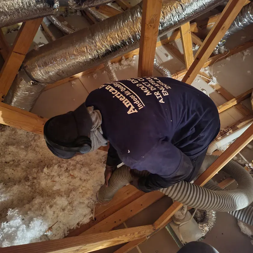 Man in an attic, installing insulation. He wears a blue shirt and protective gear.