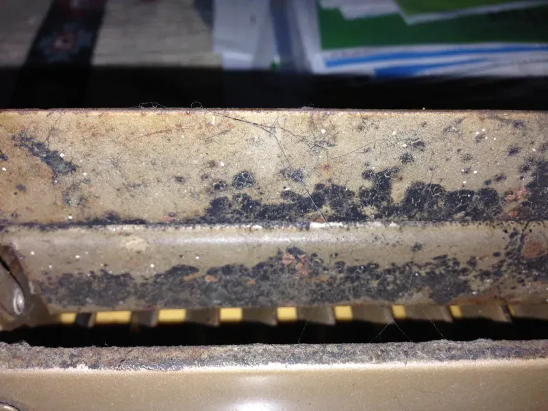 Close-up of a dirty, metal air vent with dark stains, dust, and debris.