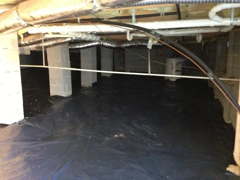 Crawlspace with black plastic ground cover, support pillars, ductwork, pipes, and wiring.