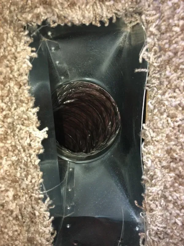 Metal air vent in carpeted floor, with a dark, cylindrical duct visible inside.