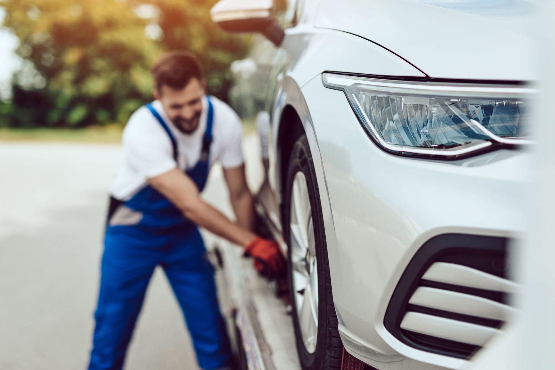 Mechanic in blue overalls inspecting the front wheel of a white car outdoors