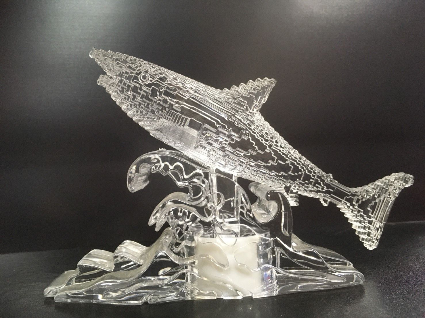 以透明玻璃製成的鯊魚雕塑，栩栩如生地展現了鯊魚躍過海浪的姿態。