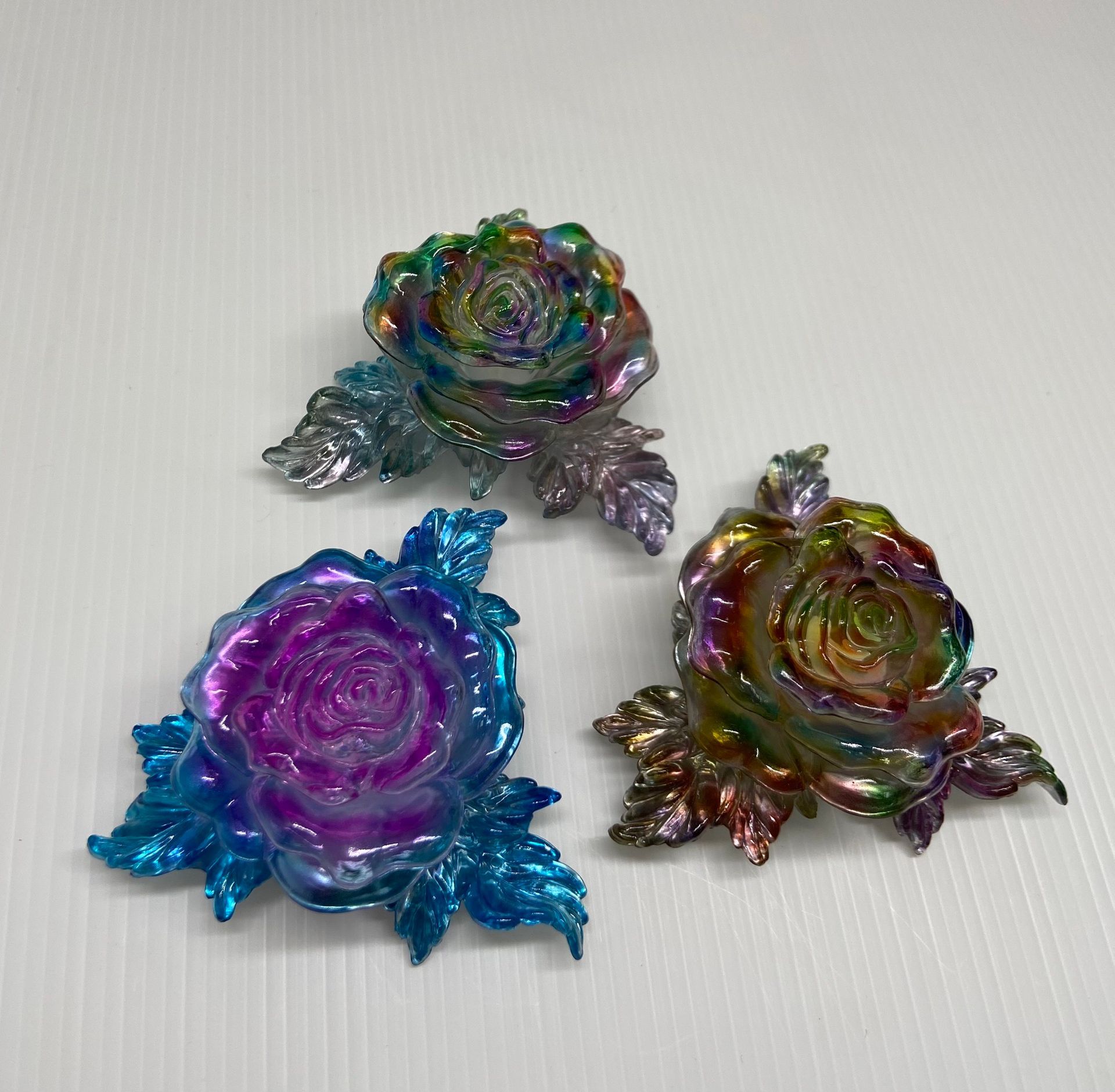 白色背景上擺放著三尊閃閃發光的玫瑰雕塑，分別呈現藍色、紫色和青銅色。塑膠飾品