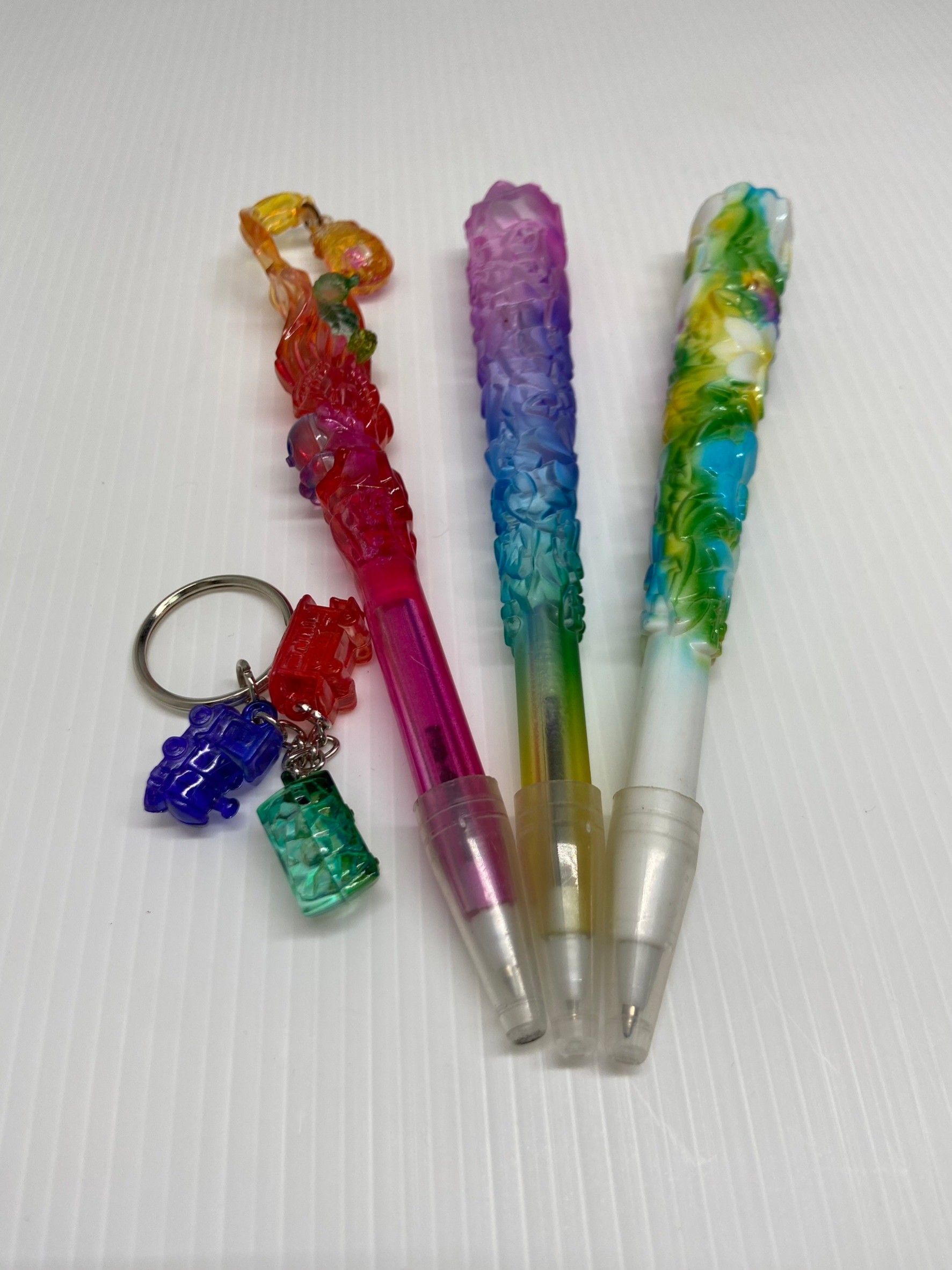白色表面上擺放著三支裝飾鋼筆和一個帶有彩色吊墜的鑰匙圈。塑膠飾品