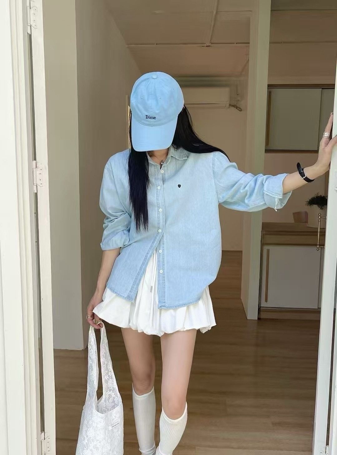 一名戴著淺藍色帽子、穿著牛仔襯衫和白色百褶裙的人站在門口，手裡拿著一個白色網袋。