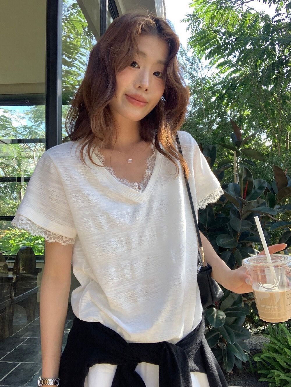一位有著棕色波浪捲髮的女士，身穿白色蕾絲邊V領上衣，腰間繫著一件黑色毛衣。