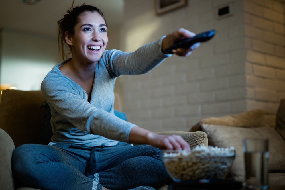 A Woman Is Sitting On A Couch Holding A Remote Control And Eating Popcorn — Craig's TV/Antenna Services In Maryland, NSW