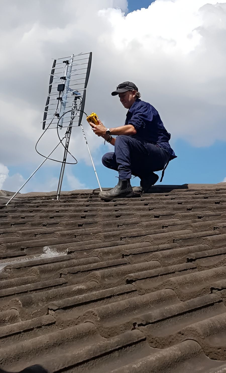 A Man Is Kneeling On Top Of A Roof Fixing An Antenna — Craig's TV/Antenna Services In Maryland,NSW
