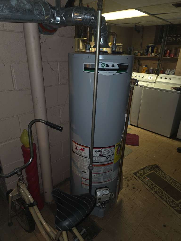 A grey water heater in a basement with a bicycle and laundry appliances in the background.