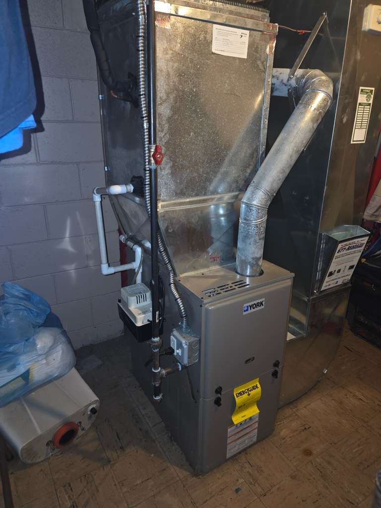 A gray HVAC furnace unit with metal ductwork stands in a room with block walls and a condensate pump on the floor.