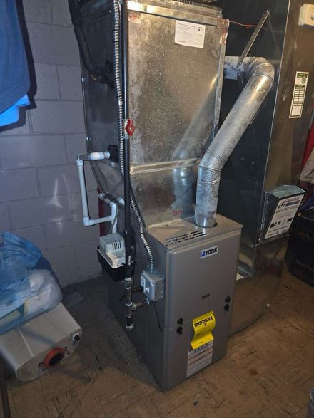 A grey furnace unit stands against a cinder block wall, featuring connected ductwork and white PVC piping.