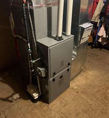 A high-efficiency furnace stands in a room, featuring dual white PVC intake and exhaust pipes and an attached drip pump.