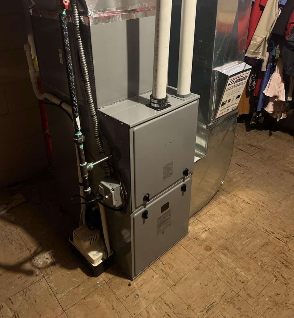 A grey furnace with two vertical white exhaust pipes stands on a wood-look floor, with clothing hanging nearby.