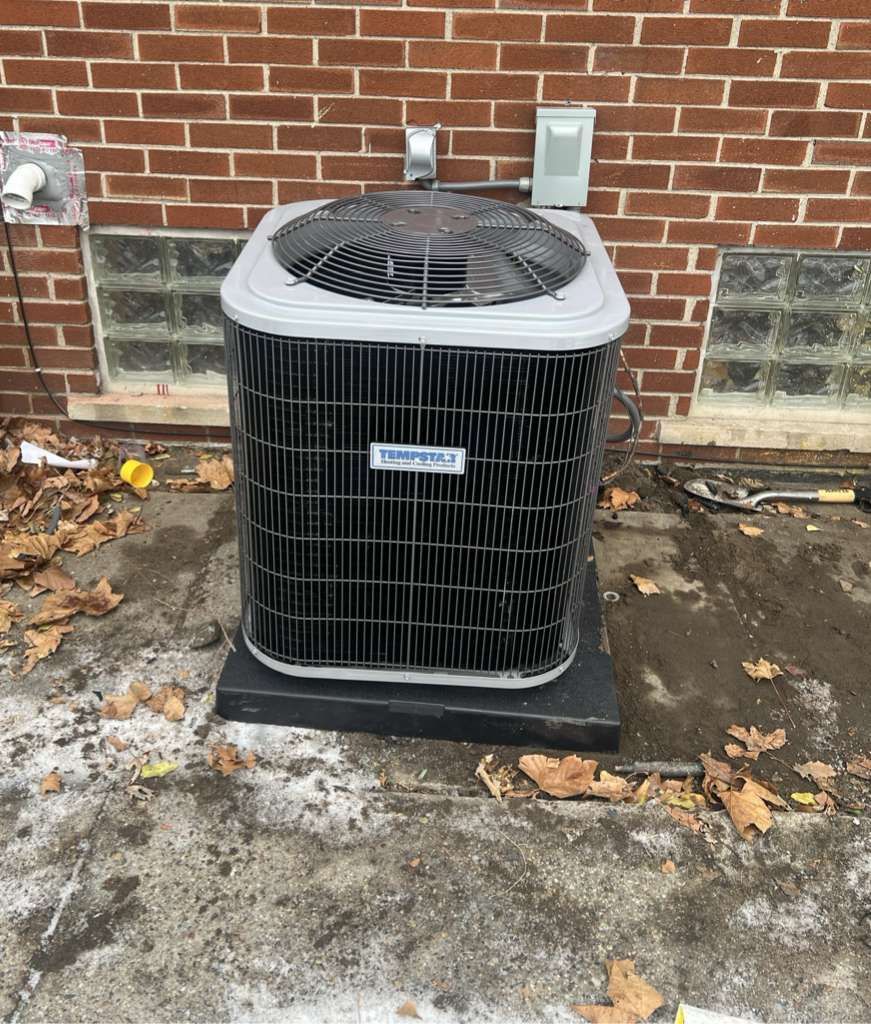 A gray outdoor central air conditioning unit sits on a black pad against a red brick wall with glass block windows.