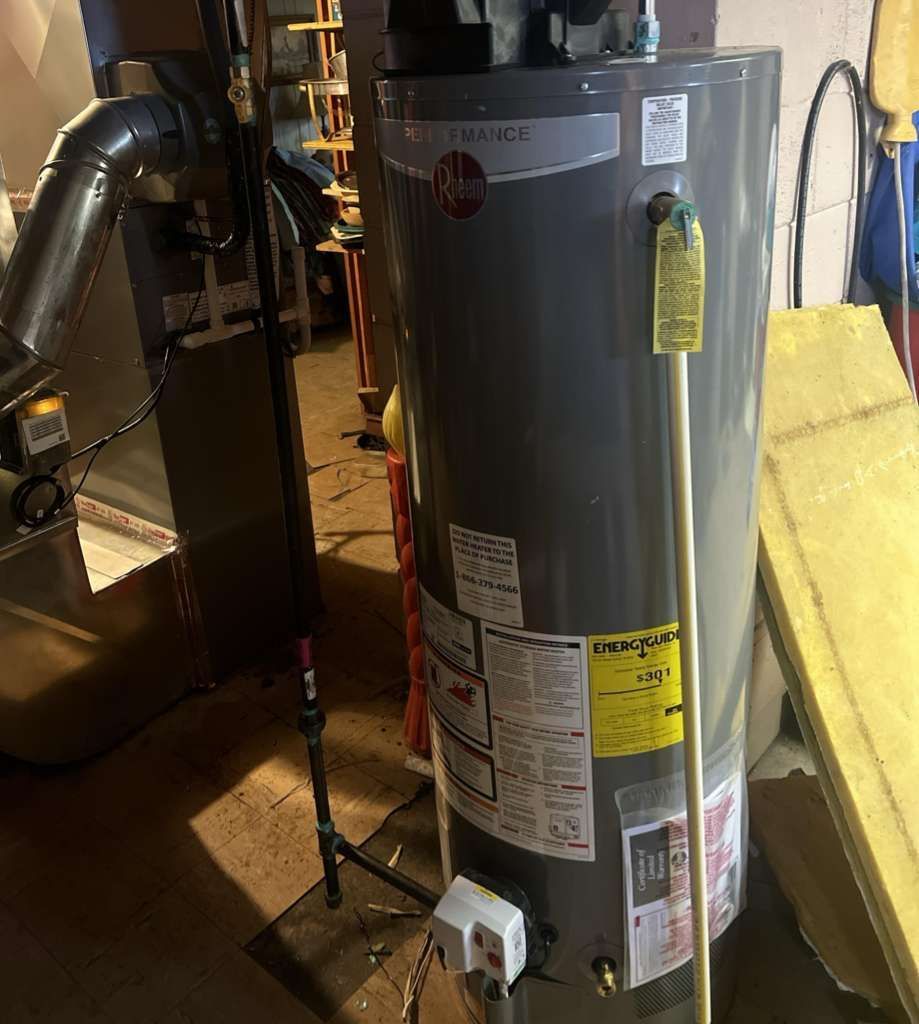 A gray Rheem water heater standing in a basement, with a yellow energy guide label attached to its side.