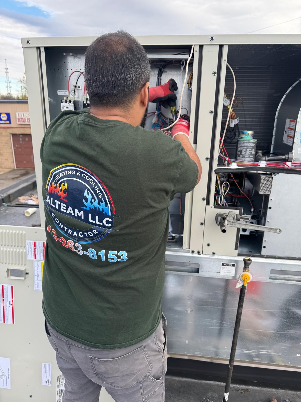 A technician wearing a dark green branded t-shirt performs maintenance on an HVAC unit on a roof.
