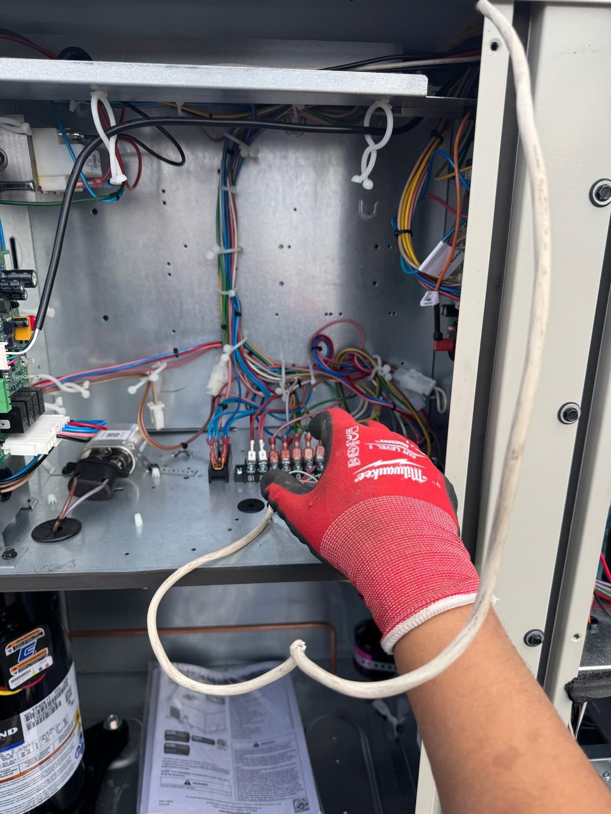 A gloved hand holds a white wire inside the open electrical cabinet of an industrial HVAC unit with visible wiring blocks.