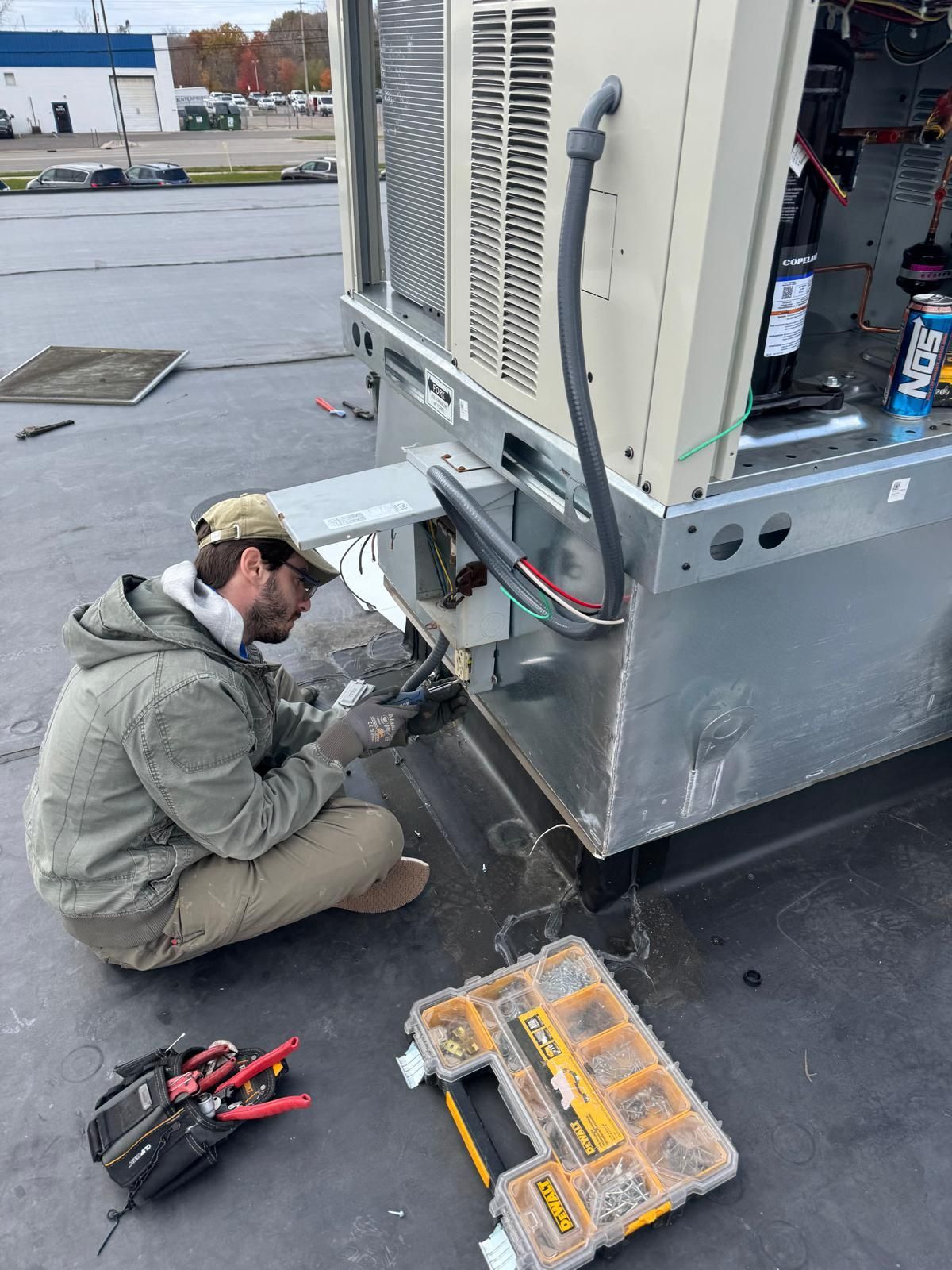 A technician wearing a jacket and cap kneels on a roof, using tools to repair an outdoor HVAC unit.