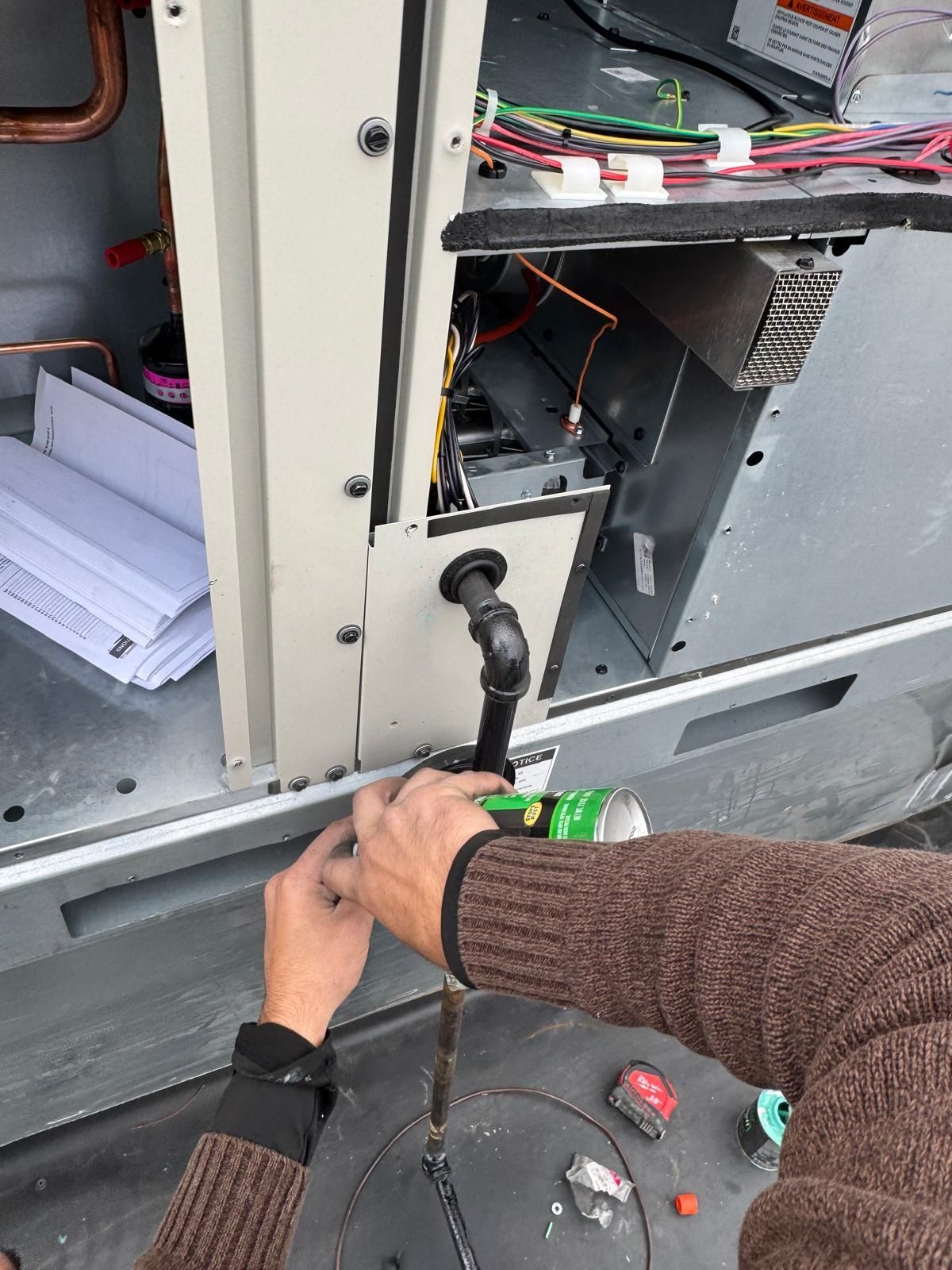 A person in a brown sweater uses a green flashlight to inspect the wiring and plumbing of an outdoor HVAC unit.