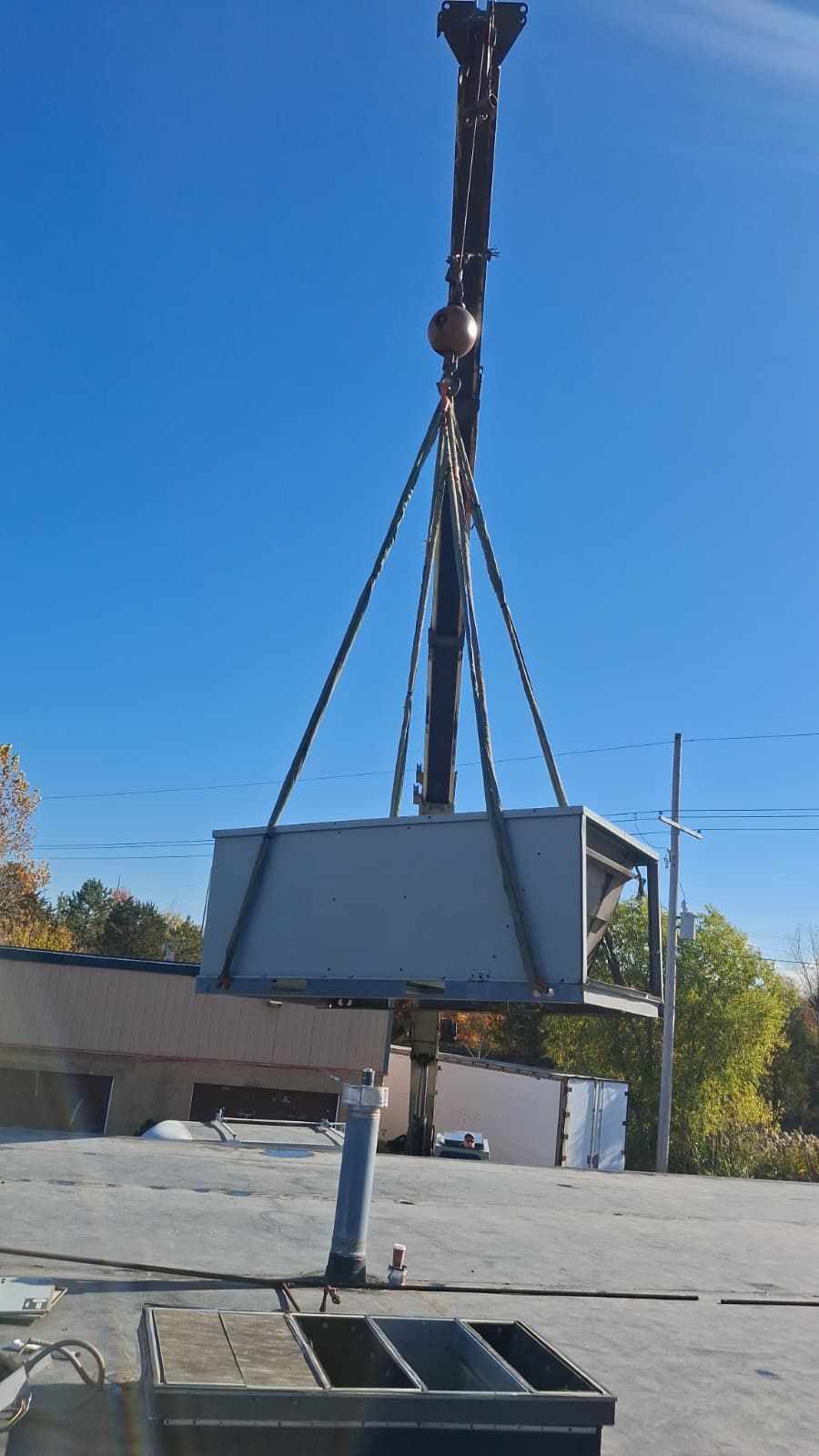 A crane suspends a large HVAC unit above a commercial rooftop on a clear, sunny day.