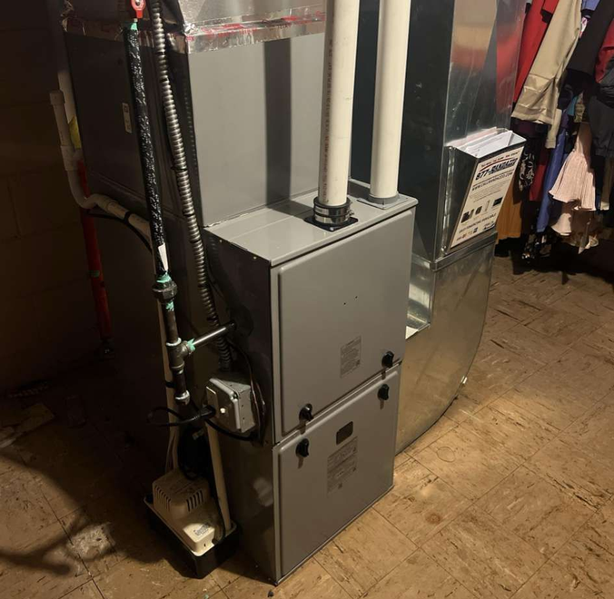 A grey residential furnace unit with white PVC venting pipes standing on a wooden floor in a utility or storage area.