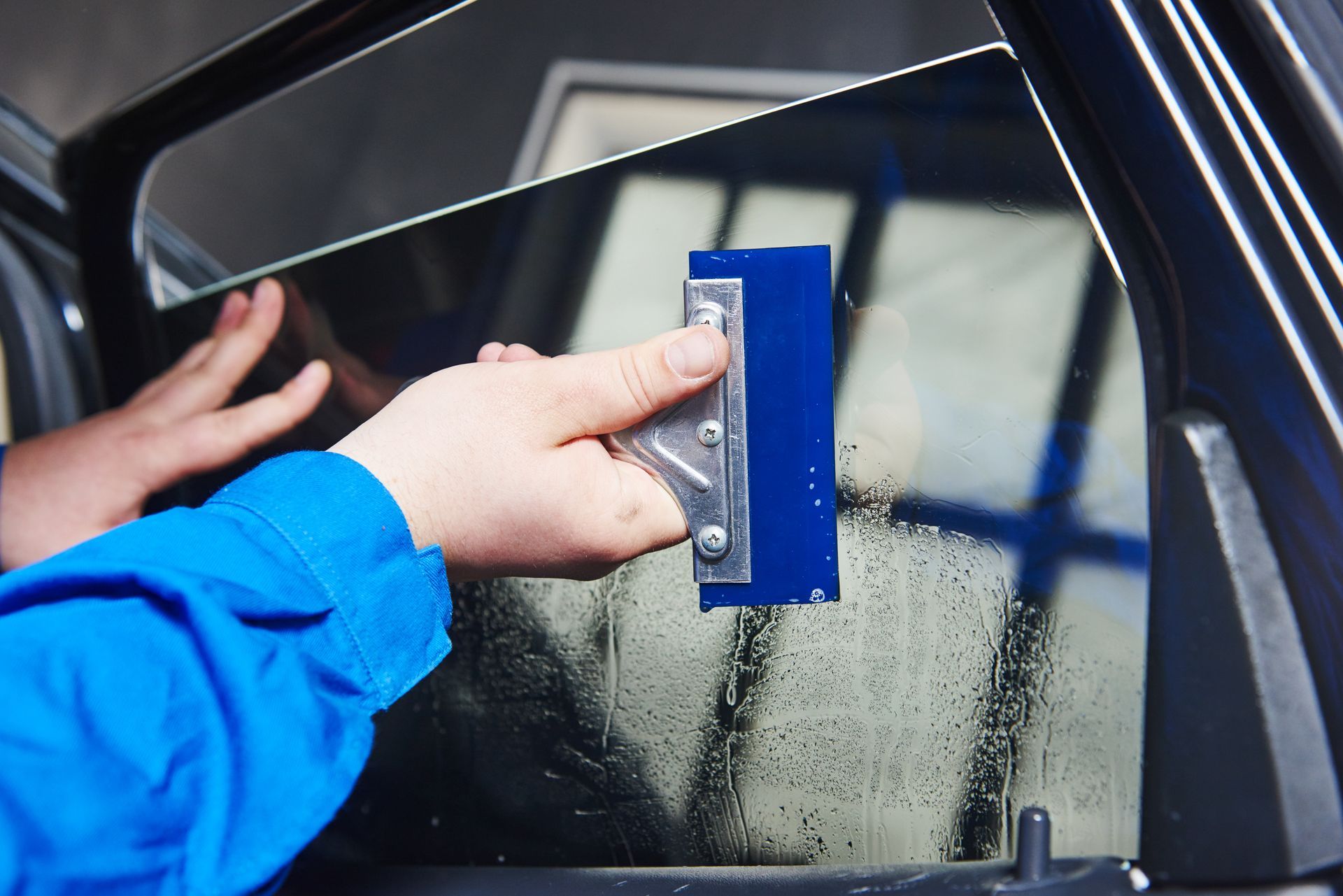 Hand using a blue squeegee to clean a car window with water droplets