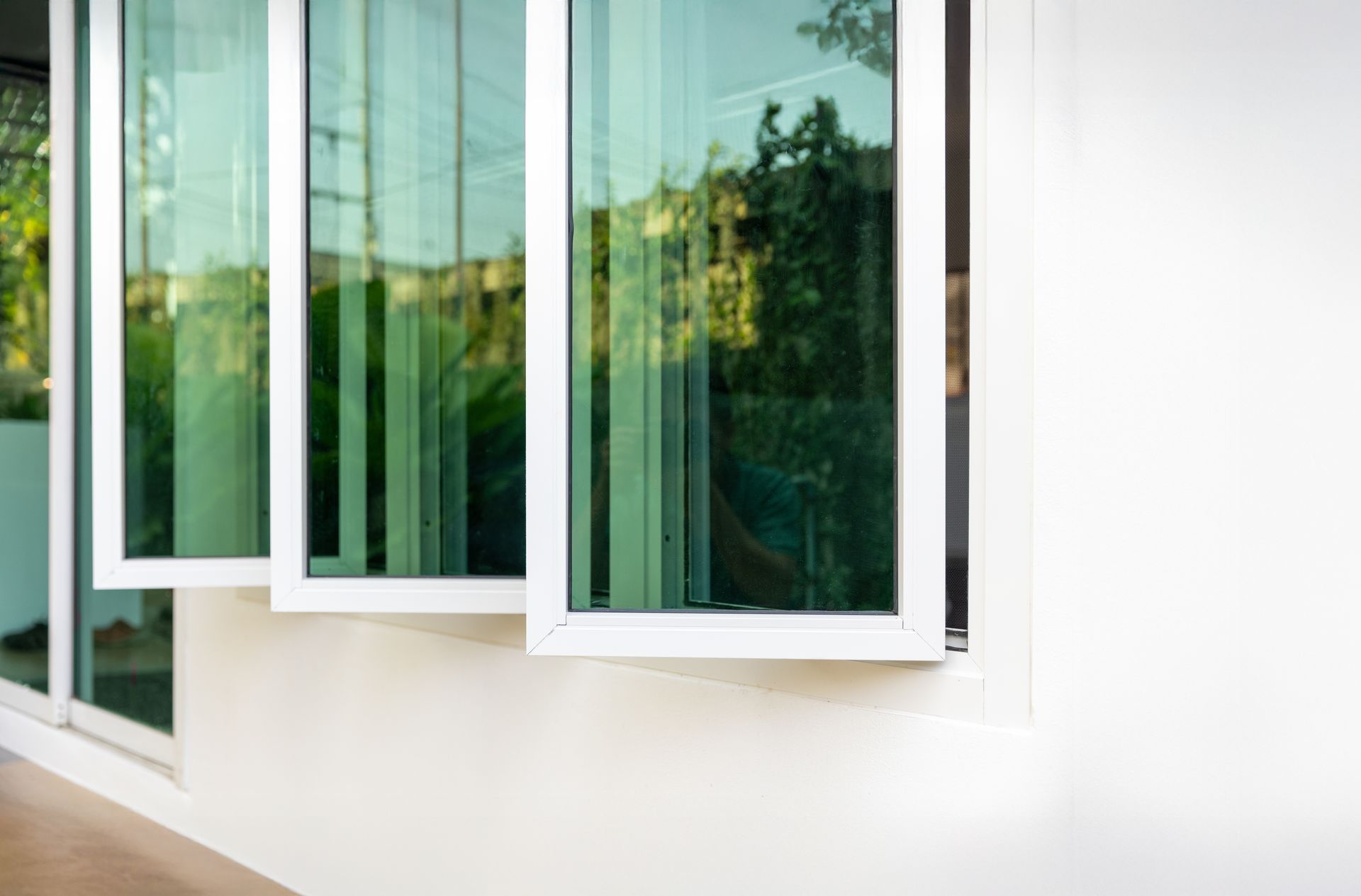 White-framed casement windows open on a modern white exterior, reflecting green trees and sky