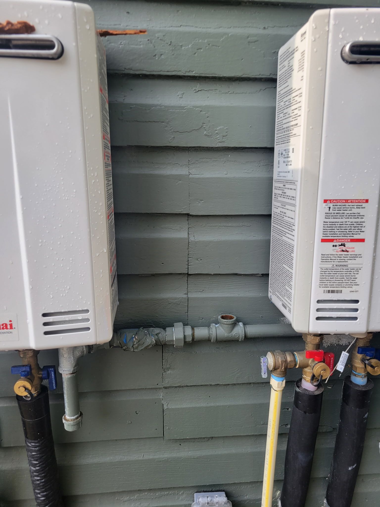 Two white tankless water heaters mounted on a weathered wooden wall, connected by a gas pipe and valves.