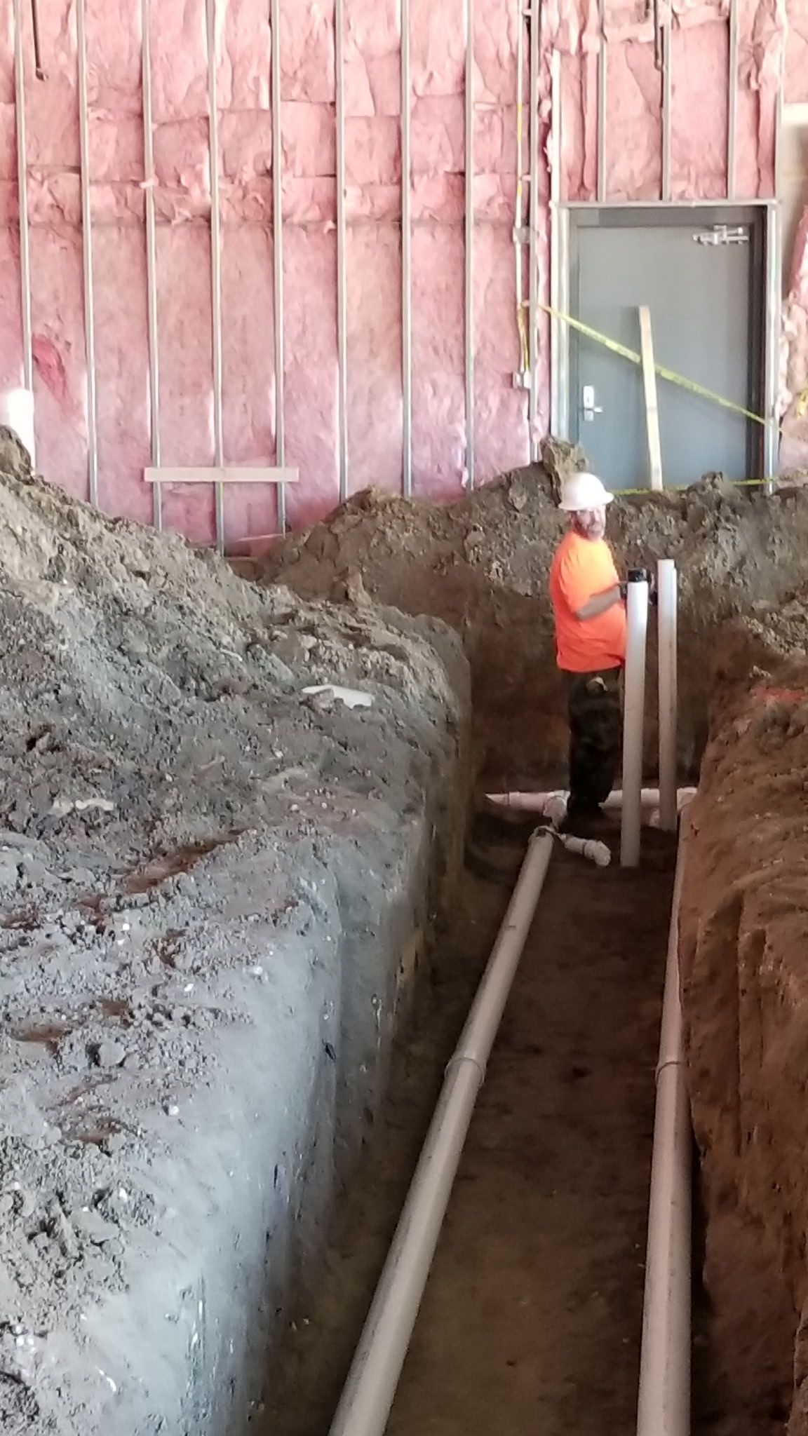 Construction worker in orange vest installing plumbing pipes in a trench at a building site.