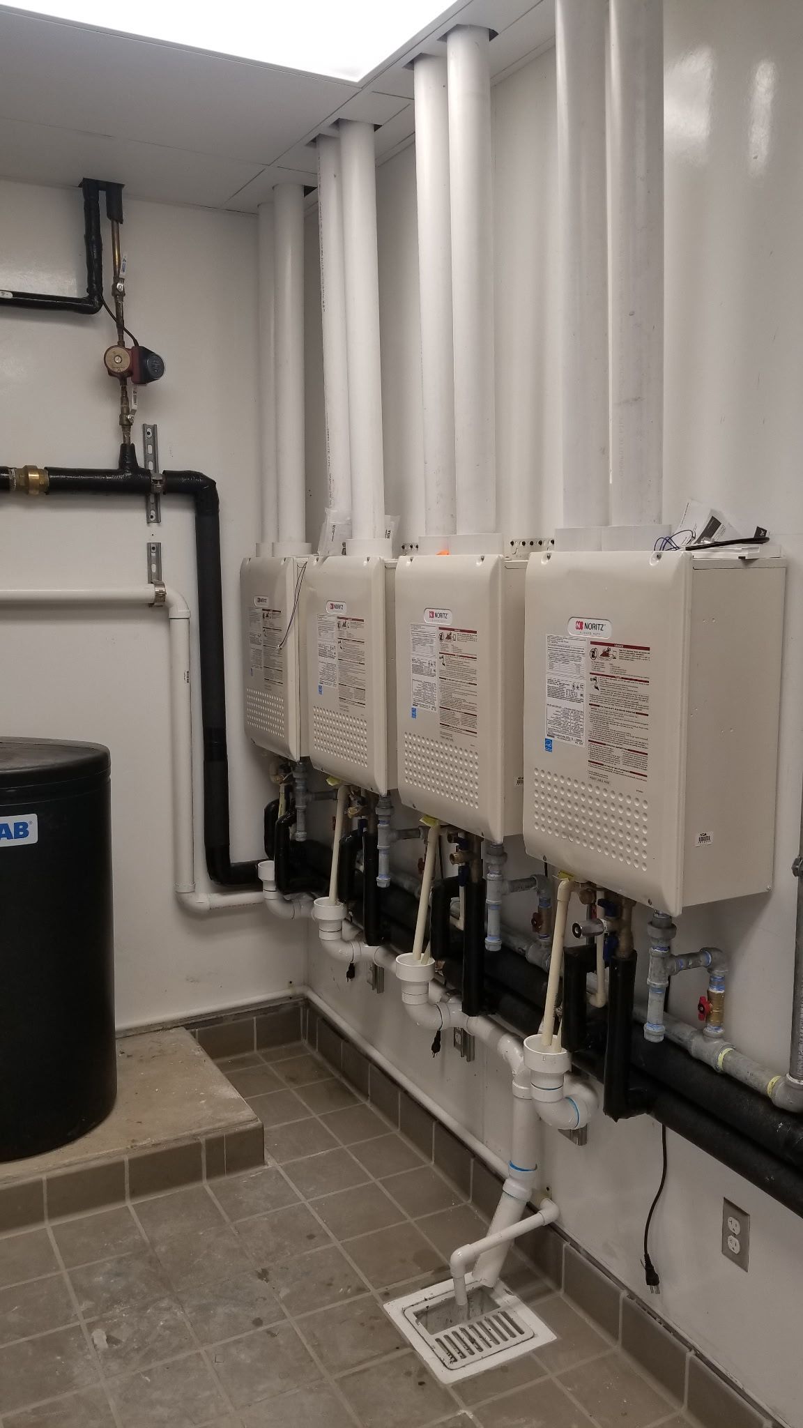 Row of wall-mounted white utility boxes with pipes in a small mechanical room.