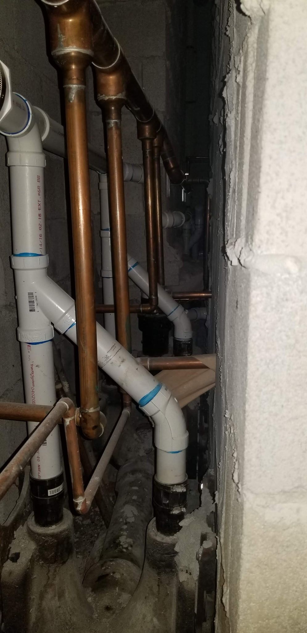 Copper pipes and white PVC plumbing running vertically in a narrow utility space against a concrete wall.