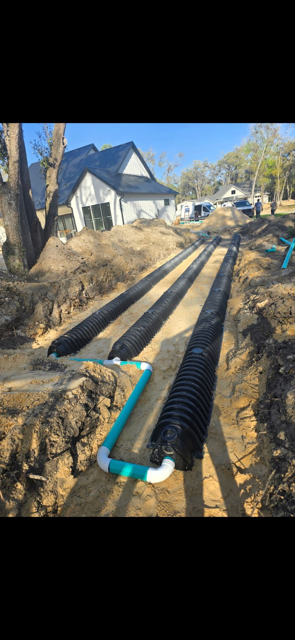 Trenches with black plastic drain pipes and white house in the background.