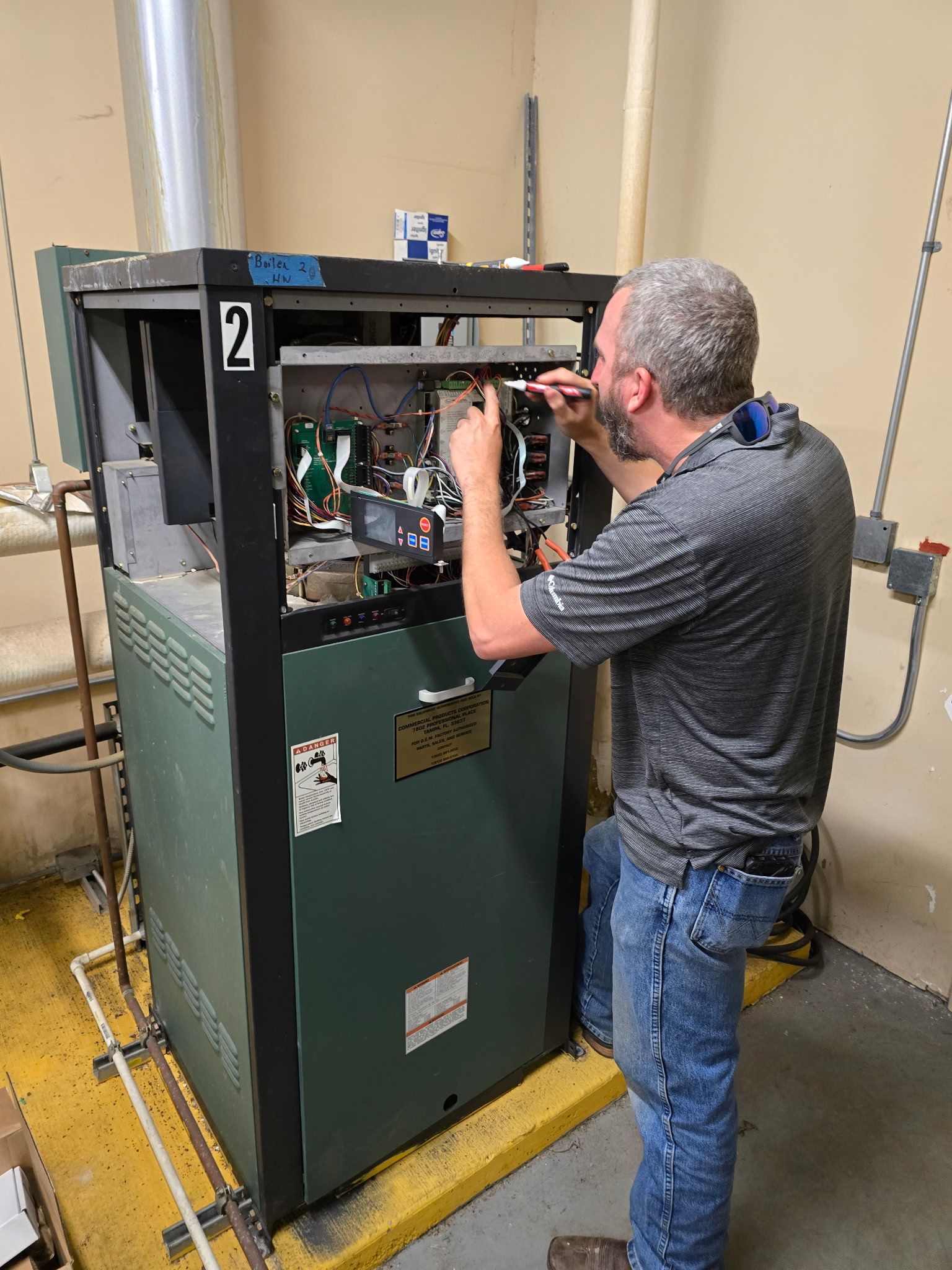 A technician in a grey shirt repairs the internal electrical components of an industrial heater unit in a utility room.
