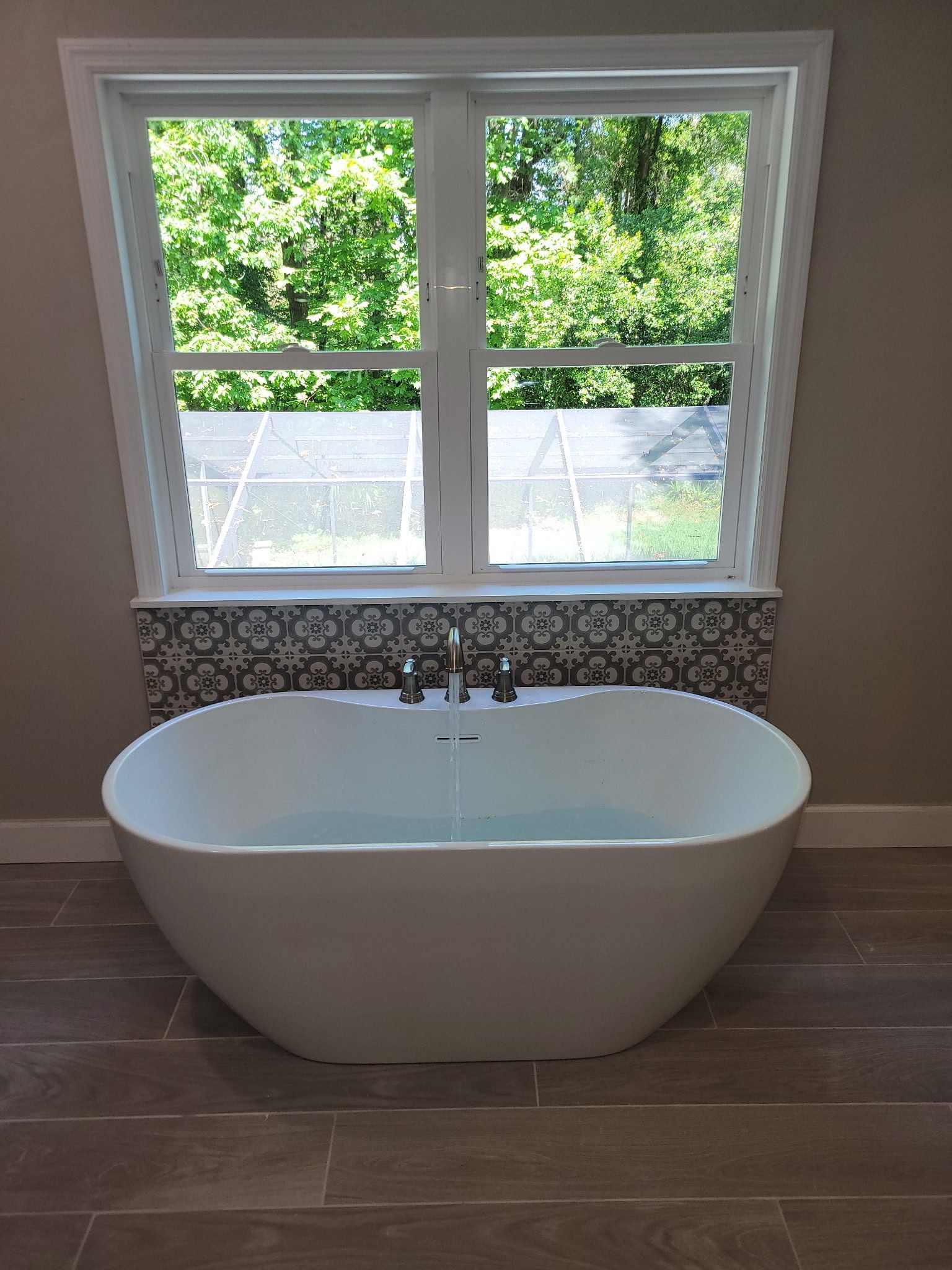Freestanding white bathtub below a window with a tile backsplash, set in a bathroom with wood-look tile flooring.