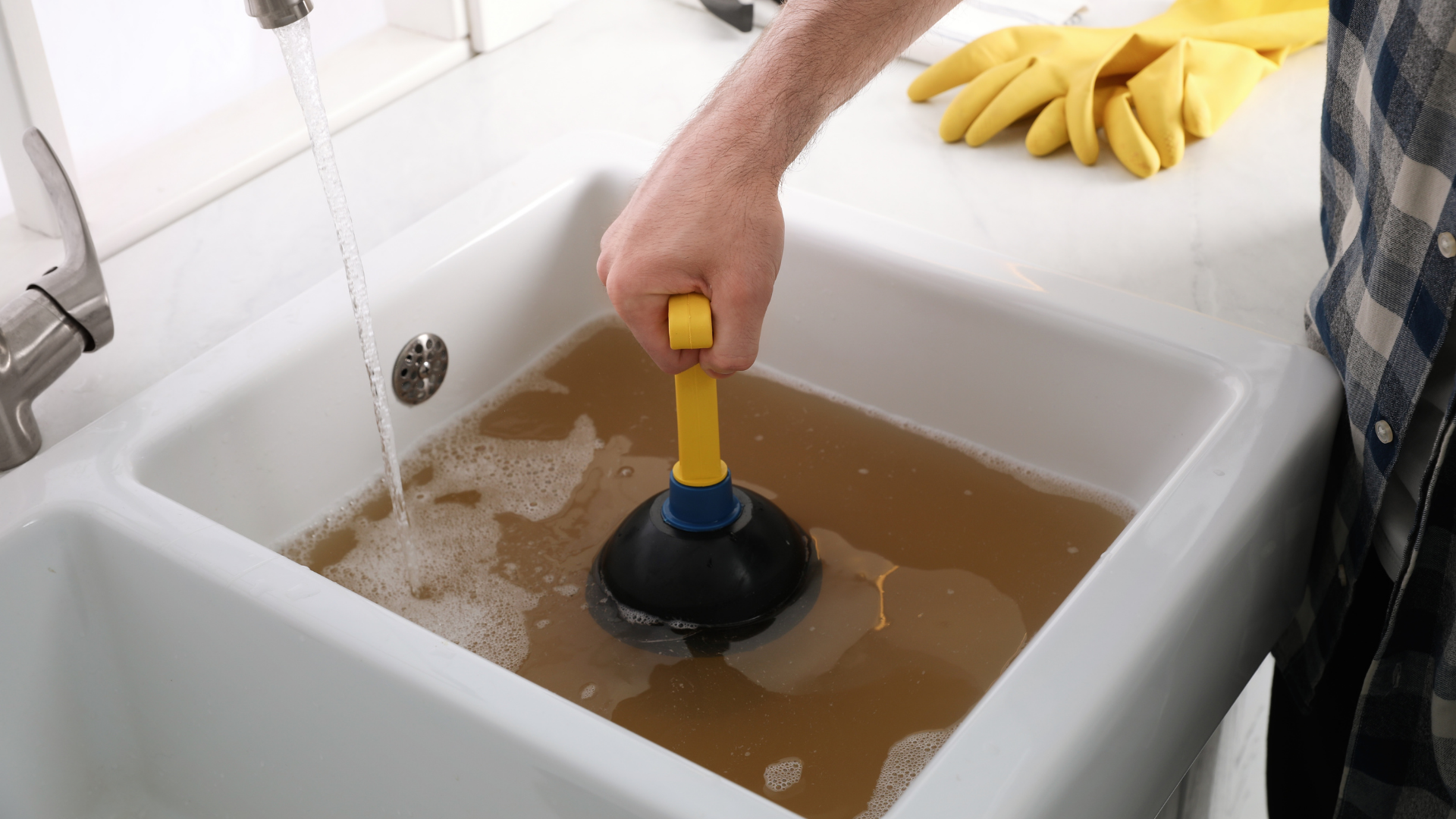 A person using a plunger to clear a clogged kitchen sink filled with murky brown water.