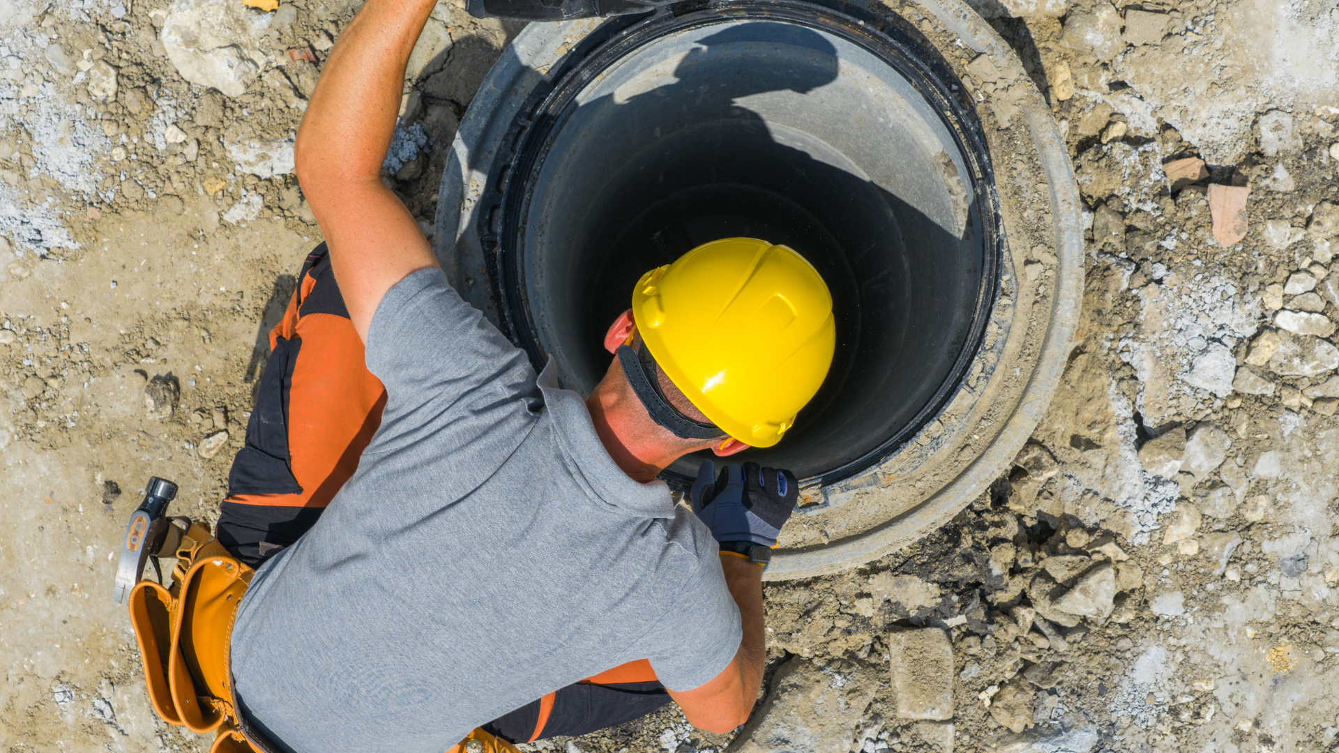 Worker in a yellow hard hat leaning into a circular manhole at a construction site