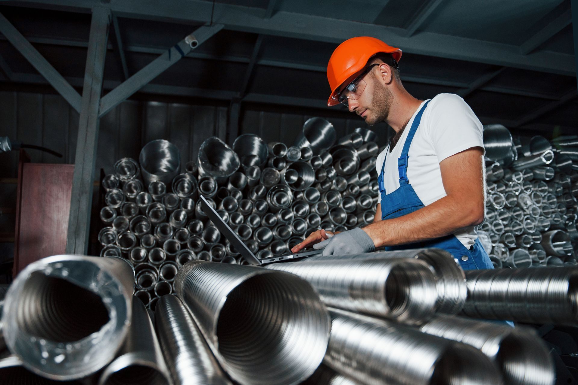 A man in work attire uses a laptop in a metal pipe factory setting A man in work attire uses a laptop in a metal pipe factory setting