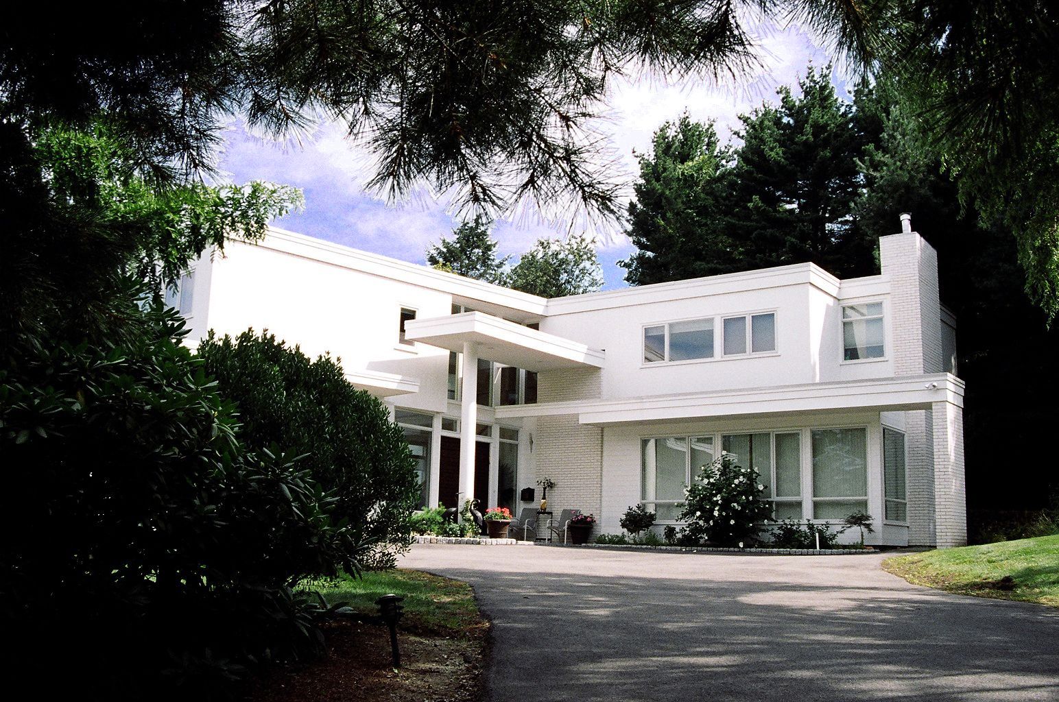 White modern house with flat roof, columns, and a driveway, surrounded by trees.