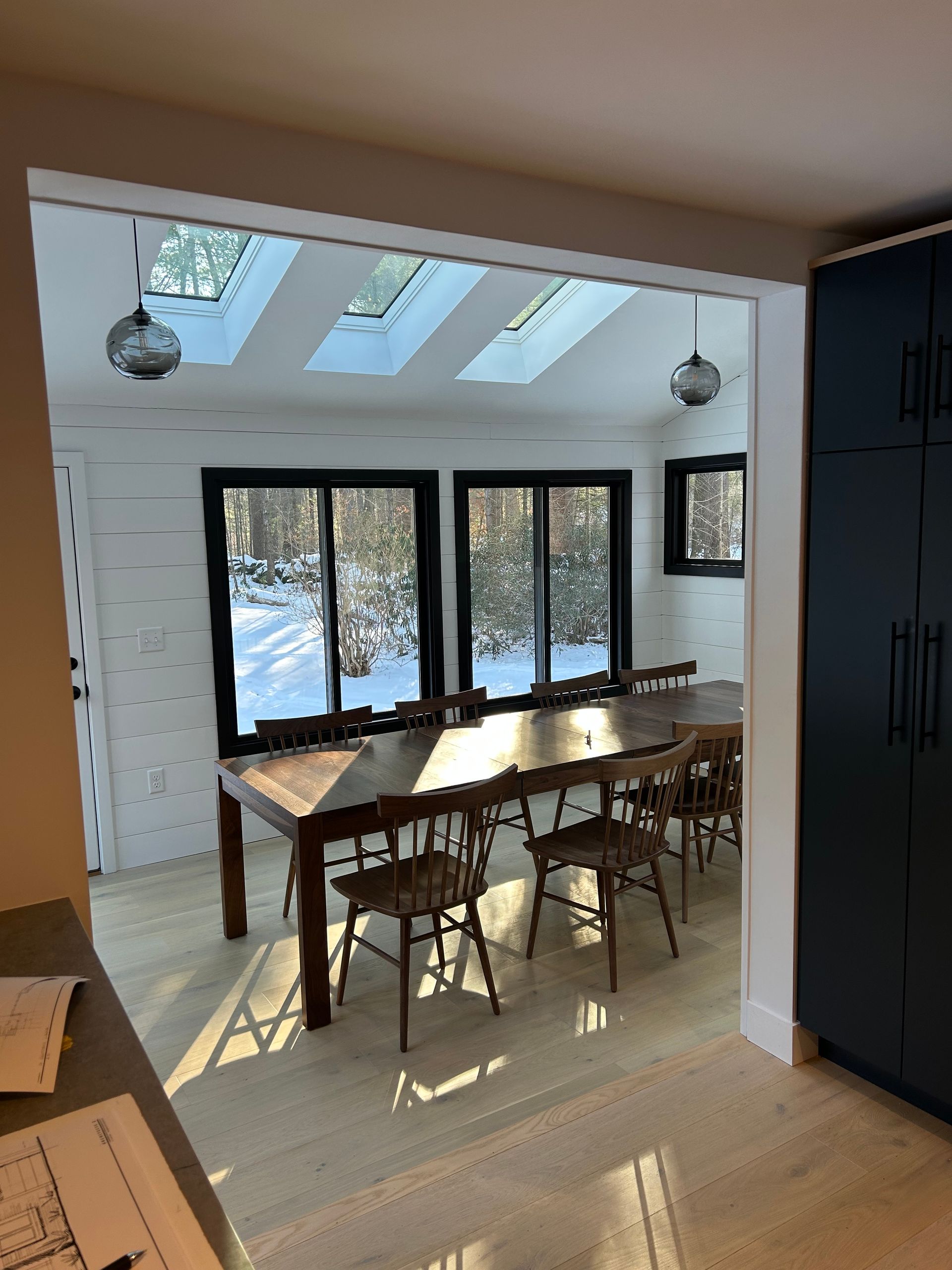 Dining room with long wooden table, chairs, skylights, windows with snow view, and black storage cabinets.