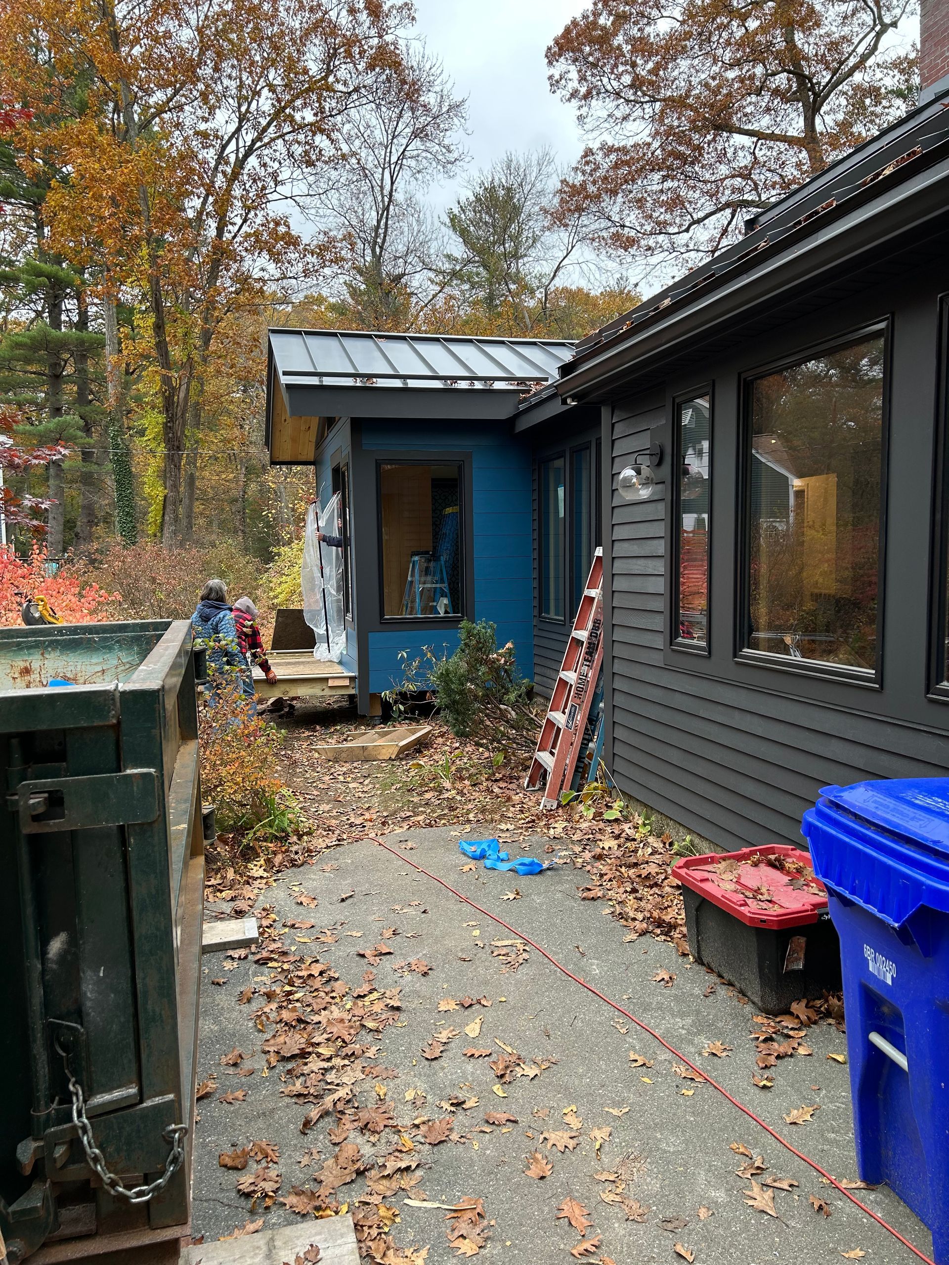 A backyard with a blue shed, gray house, dumpster, and fallen leaves.