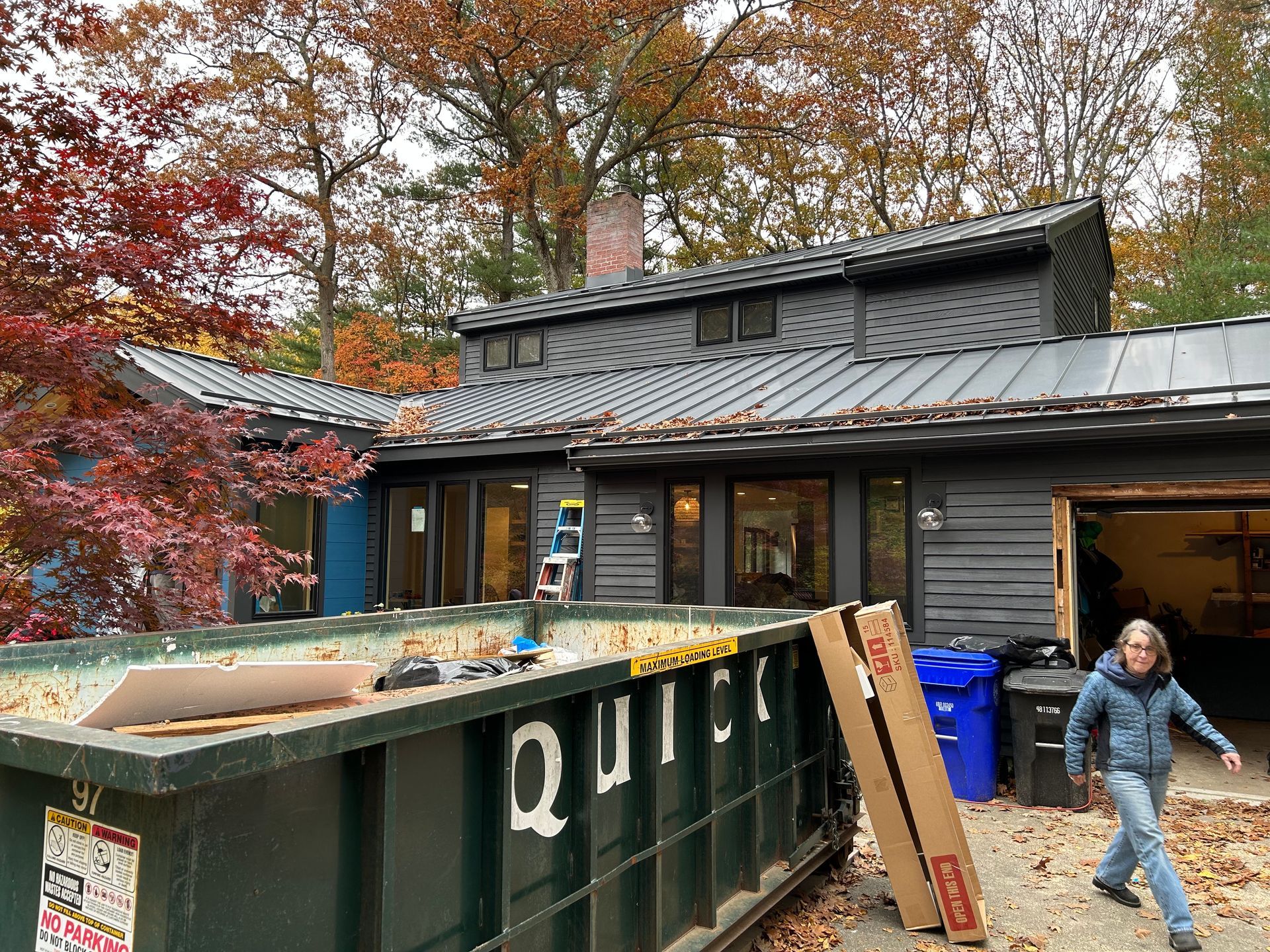 House exterior with dumpster, construction materials, and person walking. Fall foliage, gray roof and walls.