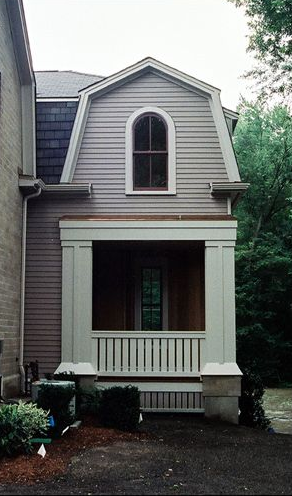 A two-story porch with a gray façade and a dark arched window. The porch has white columns and a railing.
